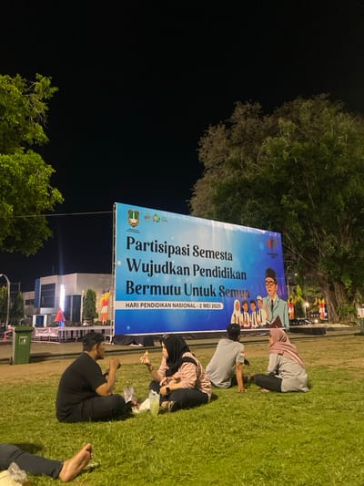 The image shows a group of four people sitting on a grassy area in front of a large billboard at night. The billboard is blue and white, with the text "Partisipasi Semesta Wujudkan Pendidikan Bermutu Untuk Semua" in large, bold letters. Below the text is the smaller text "HARI PENDIDIKAN NASIONAL - 2 MEI 2023." The billboard features a group of smiling people, some of whom are wearing traditional Indonesian clothing. The four individuals seated on the grass are all wearing casual clothing, and two of them are looking at the billboard, while two others are looking at each other. The location is Surakarta, Indonesia, at the coordinates -7.569 latitude and 110.83 longitude. The background consists of a dark sky, lush foliage, and a nearby building.