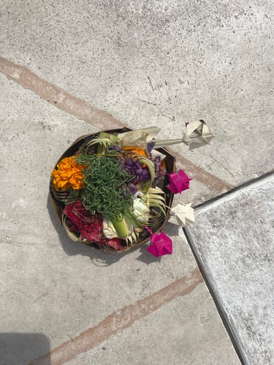 In Nusa Penida, Indonesia, an overhead shot captures a decorative basket brimming with colorful flowers and green foliage resting on a textured concrete surface. The basket, made of woven material, contains vibrant orange and red flowers, delicate purple petals, and a lush cluster of green grass. Folded paper decorations in pink and white protrude from the arrangement. The concrete ground is divided by a reddish-brown stripe, suggesting an outdoor path or patio. The scene is bathed in natural light, casting subtle shadows on the ground.