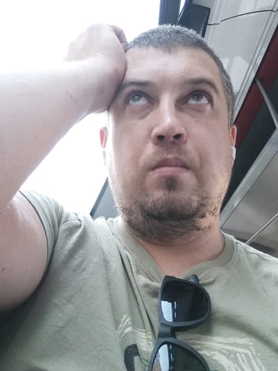 A man is leaning his head on his hand and looking up. He is wearing a light green t-shirt and sunglasses are clipped to his shirt. There is a hint of a reflection of buildings in the sunglasses lenses. The background appears to be indoors with some sort of white, metallic structure and a dark panel visible. The lighting is bright, suggesting it might be daytime. There is no indication of the location being Prague, Czechia.