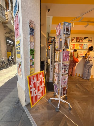 This is a frontal, eye-level medium shot of a street-facing shop entrance in Tossa de Mar, Spain. The shop's interior features a yellow and white striped ceiling. Outside, a white pillar displays several framed postcards and a vertical rack of smaller cards. A sign with red and white stripes and the words "JADE DE TOSA" in large white letters is positioned on the sidewalk. A bicycle is parked to the left of the pillar. Further inside the shop, a display rack holds various postcards, some of which have text. Two women are visible inside the shop; one is facing away from the camera, wearing a light-colored top and patterned pants, and the other is partially visible from the side, wearing a striped top and pants. Shelves with framed artwork are mounted on the back wall.