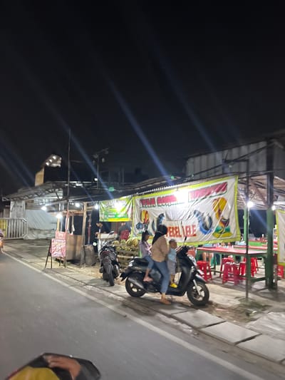 The image depicts a nighttime scene in South Tangerang, Indonesia, likely on a street or sidewalk. The primary subjects are people and motorcycles, engaged in various activities. In the foreground, a person is seated on a motorcycle. Behind them, a banner with colorful illustrations and text is prominently displayed, likely advertising a food stall or business. Several red chairs are visible, suggesting a seating area for customers.
The lighting indicates it's nighttime, with artificial lights illuminating the scene and casting shadows. The overall atmosphere appears to be that of a bustling street market or food vendor area, common in many parts of Indonesia. The presence of multiple motorcycles suggests transportation and perhaps a gathering of people.
One of the banners, though partially obscured and difficult to read completely due to the image's angle and resolution, appears to contain Indonesian text. Phrases like "AYAM GORENG" and "PECEL LELE" might be discernible, which are popular Indonesian dishes. This reinforces the idea of a food establishment.
Other details include what appear to be some stacked goods, possibly produce like coconuts, and the general structure of outdoor stalls or shops. The weather appears to be clear, as there are no visible signs of rain. The scene captures a slice of everyday life in an Indonesian urban or semi-urban environment at night.