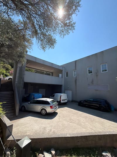 This image depicts an outdoor scene in what appears to be a residential or commercial complex, with a modern architectural style. The primary subjects are vehicles, specifically a dark-colored car parked in front of a building and a white van and a silver car parked on a concrete driveway. The building itself is gray with several windows and what looks like a ventilation grate. Trees with dense foliage are visible on the left side of the image, with the sun shining through them, suggesting it is daytime. The sky is clear and blue.

The perspective of the photo is from a slightly elevated position, looking down at the scene. There are no people visible in the image. The environment seems to be a mix of urban and natural elements, with the concrete structures contrasting with the greenery. The lighting and shadows suggest a bright, sunny day, possibly late morning or early afternoon. The presence of multiple vehicles indicates this could be a parking area or a street.

On the white van, some text is partially visible, appearing to be "kotic". The overall mood of the image is neutral and observational. The setting is likely a developed area with some vegetation, possibly in a Mediterranean climate given the type of trees. The clear sky and bright sunlight suggest fair weather. The image is oriented vertically, with the sky and trees on the left and the building and vehicles on the right.