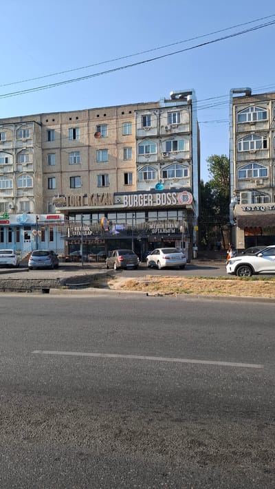 The image shows a street scene in the city of Shymkent, Kazakhstan, with a focus on a building housing a "TORT KAZAN & BURGER BOSS" establishment. The building is a multi-story apartment complex with a commercial ground floor. Several cars are parked on the street in front of the businesses. The weather appears to be clear and sunny. The architecture is typical of Soviet-era construction. The ground floor features glass storefronts and signage in what appears to be Cyrillic or a similar alphabet. One car is a white SUV parked on the right side of the street. Other cars visible are a silver sedan, a dark gray sedan, and a light gray hatchback. A person is seen walking on the sidewalk to the right of the Burger Boss establishment. A blue pharmacy with a red cross emblem is visible to the left of Burger Boss. The sky is clear blue with visible power lines overhead. The street in the foreground is asphalt with a painted white line.