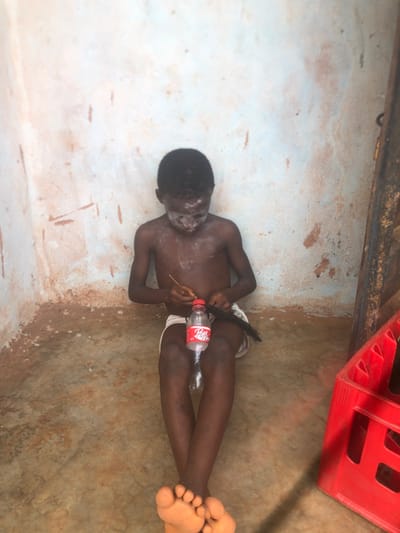 Inside a dilapidated structure in Ukwu-Abwa, Nigeria, a young, dark-skinned male child is seated on a mottled brown floor, facing forward with his gaze directed downwards. He is shirtless, wearing light-colored shorts, and has a white powdery substance applied to his face and upper chest. His bare feet are visible, crossed at the ankles. Between his legs rests a clear plastic Coca-Cola "Pop" bottle with a red cap, appearing to be cut or modified at the base. The child uses a thin, dark stick-like object in his right hand to manipulate a dark, fibrous material held in his left hand, positioned near the modified bottle. The background consists of a worn wall, painted with light blue and white patches over a reddish-brown base, showing significant discoloration and peeling, especially near the floor line. To the right of the child, a portion of a red plastic crate, containing dark bottles, is visible.