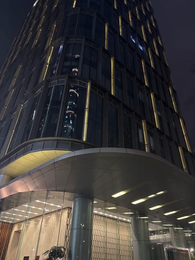 This is a low-angle, night-time photograph of the Haizhu District, China. The image features a modern skyscraper with a dark, glass facade. Vertical yellow lights accentuate the building's structure, creating a dramatic effect against the dark night sky. The lower part of the building has a glass-enclosed entrance area, illuminated by horizontal strip lighting. Reflections of other illuminated buildings are visible in the skyscraper's windows. The scene is devoid of people.