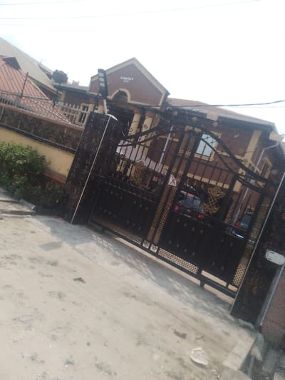 The image depicts the exterior of a property in Iranla, Nigeria, captured from an unpaved dirt road surface under daylight conditions. The scene is dominated by a large, ornate, double-leaf metal gate, primarily dark in color with gold-colored decorative elements. The gate features solid panels at its lower section and intricate scrollwork and decorative motifs in its upper half. It is flanked by dark, patterned gate posts. A small, triangular red and white sign is affixed to the right leaf of the gate.
Behind the gate, a multi-story building is visible, characterized by a prominent gabled section at the front, painted dark reddish-brown with lighter trim. The words "BY HIS GRACE HOTEL" are legible in light lettering on this gable. The building's facade is dark reddish-brown with lighter, possibly cream-colored, architectural details such as columns or pilasters. Windows with dark frames are visible. A dark-colored vehicle is partially discernible parked inside the property, beyond the gate. The roof appears to be tiled in a reddish-brown color.
To the left of the gate, a perimeter wall constructed of dark, possibly stone or concrete blocks, extends, topped with a dark barrier. Green shrubbery with scattered red flowers grows along the base of this wall. A black solar street light is mounted on a pole, positioned to the left of the gate structure and extending upwards. Overhead, utility wires traverse the upper portion of the frame against a bright, hazy sky. The foreground road surface is light-colored dirt with minor debris. No individuals are visible in the scene.