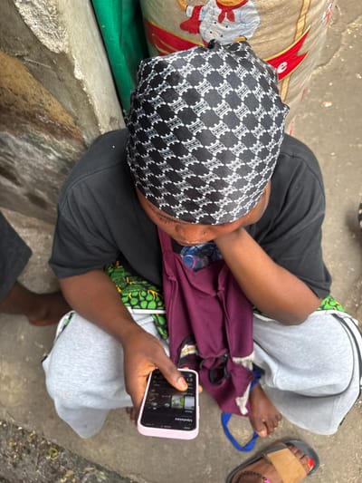A person sits on the ground, wearing a patterned head covering, a black t-shirt, and gray sweatpants. They are holding a smartphone with a pink case. A maroon shirt is partially visible on their lap. A sandal with a red-painted toe is seen near the right edge. There is a patterned bag to the left. The ground is concrete and somewhat dirty.