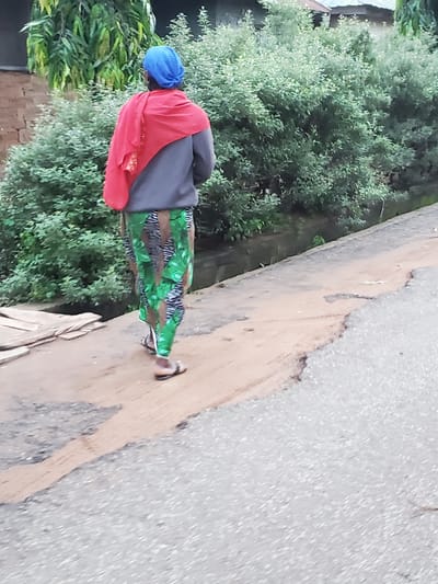 The image displays an individual walking away from the viewer on a reddish-brown dirt path in Mararaba, Nigeria. The person is dressed in a blue head covering, a grey long-sleeved top, and a red cloth draped over their left shoulder and back. They wear green, black, and brown patterned trousers and dark sandals. To the right of the dirt path is a grey asphalt road exhibiting signs of erosion and patches of lighter debris. On the far left of the dirt path, a stack of wooden planks is visible. The background consists of dense green foliage, including tall leafy trees on the left and a cluster of light green bushes in the center-right. Above the bushes, a section of a building's roof, possibly corrugated metal, is partially visible.