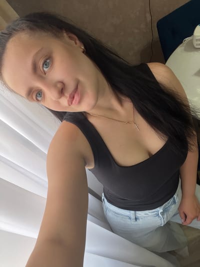 An indoor selfie captures a young woman with long, straight dark hair and light blue eyes. She is wearing a black sleeveless top and light-wash blue jeans. A delicate gold chain necklace with a small, sparkling cross pendant is visible around her neck. The woman looks directly at the camera with a calm, neutral expression, her head tilted slightly. Her arm is partially visible on the right side of the frame, suggesting she is holding the camera.
The background on the left shows a dark brown floor with a black cord and parts of white and dark blue objects, possibly furniture or bedding. On the right, bright, diffused light filters through white, translucent curtains, indicating a window and suggesting it is daytime. There is no visible text in the image.