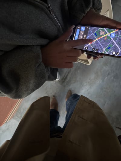 A close-up overhead shot depicts a person holding a smartphone displaying a Google Maps navigation interface, likely in Yerevan, Armenia, but NOT in the city of Zovuni, Armenia. The screen shows directions indicating a 24-minute, 8-kilometer route with Yerevan Opera House visible as a landmark. The person's hand is touching the screen, apparently interacting with the map. The person is wearing a grey zipped jacket and a white bag is seen partially. Another person wearing blue jeans, and brown shoes and a light tan coat is partially visible on the right side of the frame. The background surface is a neutral grey.