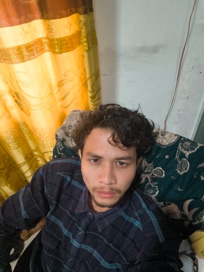 In Pulodolang, Indonesia, a person with dark, curly hair and a light complexion is captured in a selfie. They are wearing a dark plaid shirt with blue and purple stripes and are seated on a patterned cushion, which appears to be part of a chair or sofa. Behind the subject, a yellow curtain with a subtle pattern is visible, partially obscuring a white wall. A thin red wire runs vertically along the wall. The lighting suggests an indoor setting.