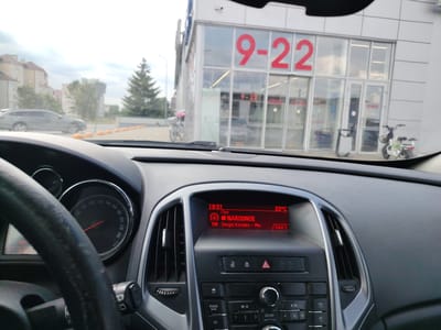The image, taken from inside a car, shows a view of a store with large red numbers "9-22" on its facade, indicating the store's operating hours. The store appears to be located in Malaryta, Belarus, as suggested by the context of the location. To the left of the store, a dark-colored station wagon is parked, and further in the background, residential buildings and a conifer tree are visible. A bicycle is parked near the store entrance. Inside the car, the dashboard is prominent, with a steering wheel on the left and the car's radio display in the center, showing the time as 18:01, temperature as 23°C, and a radio station playing "NARODNOE" with the artist listed as "Sergej Kovalev - Min." A white electric scooter is parked to the right of the store entrance. The overall scene suggests a daytime setting in an urban or suburban environment.