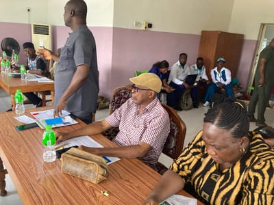 An indoor scene, likely a meeting or training session, is depicted in Umuahia, Nigeria. The room features light-colored walls, combining white and a pale purplish-pink hue. Wooden tables are arranged with chairs for participants.
In the foreground, a man in a dark grey short-sleeved top stands, gesturing with his right hand as if speaking or presenting to the group. Seated at a wooden table in front of him, an older man in a red and white plaid shirt, a light brown cap, and sunglasses looks attentively towards the speaker. His hands rest on papers spread across the table. Beside him, a woman in a distinctive yellow and black zebra-print blouse with braided hair is engrossed in reviewing documents on the table.
Further down the table, numerous clear plastic water bottles with green caps (some showing the "Aquafina" brand) are distributed. Papers, pens, and mobile phones (including one with a blue case) are also visible on the tables.
In the mid-ground and background, other attendees are seated. An older man in a black cap and patterned shirt is visible at another table. Several younger individuals are seated against the far wall; some are using laptops or smartphones, while others are observing. One woman in a blue top and trousers wears a light blue baseball cap with partially visible text "O Pa" or "Opo".
The room is equipped with a standing black fan on the left and a wall-mounted air conditioner unit. A large wooden wardrobe or cabinet stands against the back wall. The lighting suggests daytime. The overall atmosphere appears focused and professional, indicative of a learning or discussion environment.