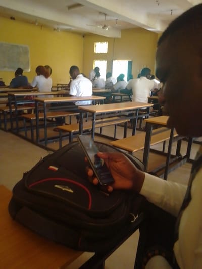 A classroom scene in Awka, Nigeria, features students seated at desks. The image foreground shows a student holding a mobile phone with a black backpack on the desk beside them. A row of occupied desks with students wearing white shirts extends into the background. The room is painted yellow with natural light coming from the windows.