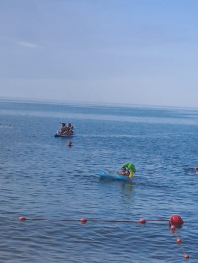 In the city of Dzhubga, Russia, on a clear day, the blue sea is calm and inviting. Several individuals are enjoying recreational activities on the water. In the distance, a person is seen swimming, while a child sits on a blue inflatable raft with a palm tree design. Closer to the foreground, two people are seated on a paddleboard, each wearing a life vest and holding a paddle. In the foreground, a rope with several red buoys stretches across the water, marking a boundary. The scene conveys a sense of leisure and outdoor enjoyment.