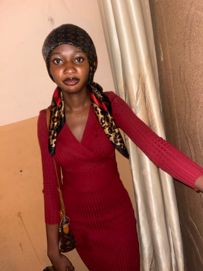 A woman is depicted indoors, possibly in a room in Unguwar Gabas, Nigeria, given the location context. She is wearing a long-sleeved, red ribbed dress and a headscarf with a black and gold patterned exterior. The headscarf is draped around her head and neck, with portions falling on her chest. She has dark hair, dark eyes, and appears to be facing the camera. A piercing is visible above her chest. In one hand, she holds a pair of glasses and their case. The background includes a beige wall and a curtain or drape.
