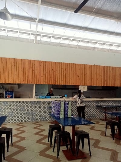 The scene shows a restaurant interior in Pati, Indonesia, located at coordinates -6.752 latitude and 111.026 longitude. Two people are present. A man wearing a white shirt and a black cap is standing behind a counter. He is facing the right side of the image. A woman wearing a light blue and white dress is standing at a counter, facing the left side of the image. She is holding a stack of paper in her left hand and reaching with her right hand toward the counter. Behind the counter are a number of boxes stacked on a table. The counter has a white countertop and a blue and white tiled backsplash. There are four tables in the scene, each with four chairs. Two of the tables have blue tops, and the other two have brown tops. All of the chairs are black and are made of metal. The restaurant has a white ceiling with a skylight. The walls are white, with a wood paneling accent. The floor is tiled with beige and brown square tiles.