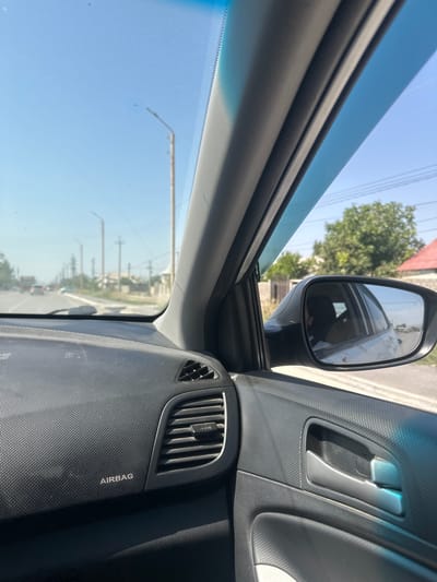 This media file captures an interior view of a car, likely in Kyrgyzstan, during daylight hours. The scene is viewed from the perspective of a passenger or driver, looking forward and to the side.

Prominent objects include the car's interior elements such as the door panel with a silver interior door handle, a dashboard vent, and a textured surface with the word "AIRBAG" printed vertically. The side mirror is visible, reflecting a portion of the car's interior and the exterior scenery. Through the side window and the windshield, a road can be seen with trees and buildings lining it, suggesting an urban or suburban environment. Overhead power lines are also visible in the sky. The sky is a clear blue, indicating fair weather.

The overall impression is that the car is either parked or moving slowly along a road. The lighting suggests it is daytime, likely mid-morning or afternoon, with bright sunlight. The image has a slightly tilted orientation, adding to the dynamic feel of being inside a moving vehicle. No people are clearly visible, though reflections in the mirror might hint at their presence. The setting appears to be a developed area with infrastructure like roads and utility lines.