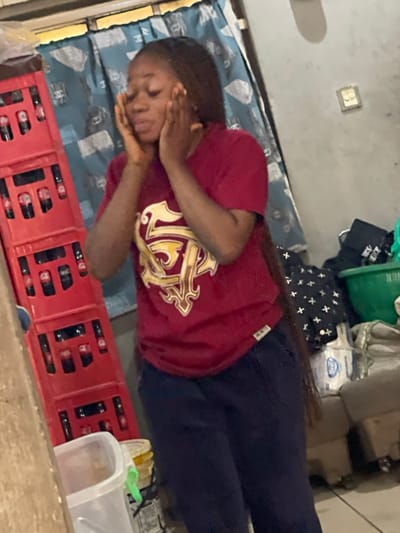 A young woman lies on a bed, her head propped on her hands. She's wearing a maroon t-shirt with a large gold crest. The bed has a blue and white patterned cover. Next to her is a red plastic crate filled with dark bottles. In the background are assorted items including a green basket, bags with white crosses, and white boxes. A light switch plate is on the wall behind the bed. She looks distressed.