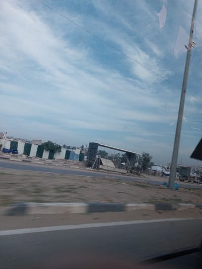 The image, captured from a moving vehicle in a-Zawayda, Palestinian Territory, depicts a roadside environment under a partly cloudy sky. The immediate foreground shows a grey asphalt road with a white lane line, topped by a concrete curb painted in alternating white and dark grey sections. Beyond the curb, a dirt and gravel shoulder extends across the frame, exhibiting motion blur. Across this shoulder lies another asphalt road. On the far side of this road, a tall, dark utility pole stands vertically on the right. To its left, a large, dark-colored metallic gantry-like structure, partially clad with corrugated metal sheeting, dominates the midground. Adjacent to this structure and extending leftward, a barrier or fence composed of alternating white and dark green/blue vertical rectangular panels is visible. Behind this barrier, various debris and rudimentary structures are scattered, along with sparse green vegetation. In the distant background on the left, indistinct urban buildings are present. The sky above is light blue with scattered white, streaky clouds, and a faint, dashed line, consistent with a reflection, traverses the upper portion of the sky. No individuals or discernible actions are depicted.
