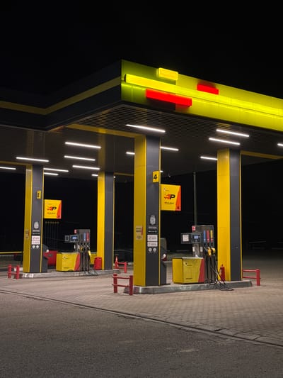 This image depicts a modern, brightly lit gas station at night, located in Maksimkovo, Russia. The station features a distinctive yellow and black canopy structure with vibrant yellow lighting and red accents along its edge. Several illuminated gas pumps are visible, each with attached hoses and nozzles. On the left side of the image, a sign with the "HP PULSAR" logo is displayed on a yellow panel. To the right of this sign, a red fire extinguisher is mounted on a fuel dispenser. The ground is paved with interlocking stones, and red bollards are placed around the fueling area. The background is dark, suggesting it is deep night. No people are visible in the frame.