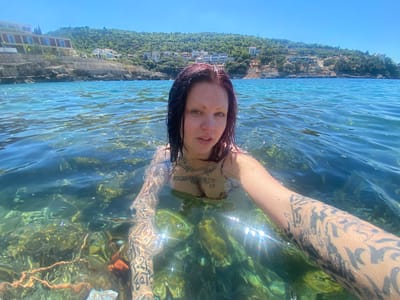 A woman with dark hair and tattoos swims in clear, turquoise water. She is holding the camera for a selfie. Her tattoos are visible on her arm and chest. The seabed is visible through the clear water, with rocks and seaweed. In the background is a hill covered in green vegetation and buildings, a seaside resort area. The location is the Adriatic Sea. The area in the city of Kunje, Montenegro. It is daytime, and the sky is clear. The weather is sunny.