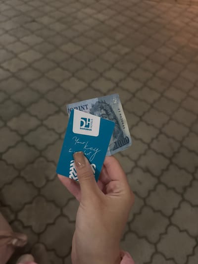 A person is holding a Hungarian 1000 Forint banknote and a blue hotel key card. The banknote has the serial number JE 8020645. The key card is for Danubius Hotels and has the text "Your key to comfort" printed on it. The background appears to be an outdoor paved area with a repeating geometric pattern. The lighting suggests it is daytime. The image is taken from a top-down perspective.