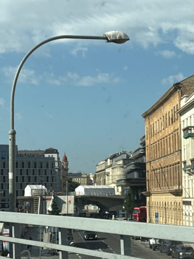 The image shows a street scene in Prague, Czechia, with a lamppost in the foreground on the left. Behind it, a row of buildings lines the street, with a mix of architectural styles. The buildings on the right are older, with ornate facades and many windows. The buildings on the left appear more modern, with a grid-like pattern of windows. In the background, a red truck is visible, along with several cars. The sky is blue with scattered clouds. The scene appears to be during the daytime, with good visibility.