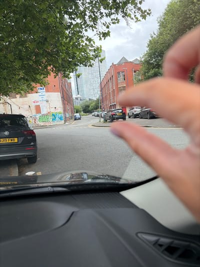The image depicts a street scene in Salford, United Kingdom. In the foreground, the dashboard of a car is visible. A hand, with painted fingernails, is positioned in the mid-ground, creating a framing effect over the scene.
The street is paved and appears to be an urban road. Several cars are parked on the side of the road and one car, a black Volkswagen Tiguan with license plate NJ19 FWB, is in the left lane, facing away from the viewer. Further down the street, other vehicles are present.
Buildings line the street. On the left, a red brick building displays advertisements, including one for "TNT Textiles & Hosiery Ltd." and a "TO LET" sign. Graffiti adorns the lower section of this building. On the right, another red brick building with multiple windows is visible. In the background, a modern, tall glass building dominates the skyline, indicating a contemporary urban setting. Trees with green foliage are present on the left and right sides of the image, contributing to the urban greenery. The sky is overcast, suggesting a cloudy day.