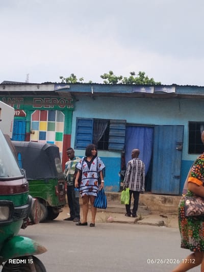 The image depicts a street scene in Aba, Nigeria, with a blue building serving as a backdrop. In front of the building, several people are visible. A woman in a patterned dress is walking away from the camera. Another man, also in a patterned shirt, is standing and looking down. To his left, a man in a plaid shirt is also standing. In the foreground, a green and maroon auto-rickshaw is parked. The building itself appears to be a shop or business, with a sign that partially reads "INT DEPOT". The street is paved, and there is some vegetation visible in the background. The overall impression is of a typical commercial area in the city.