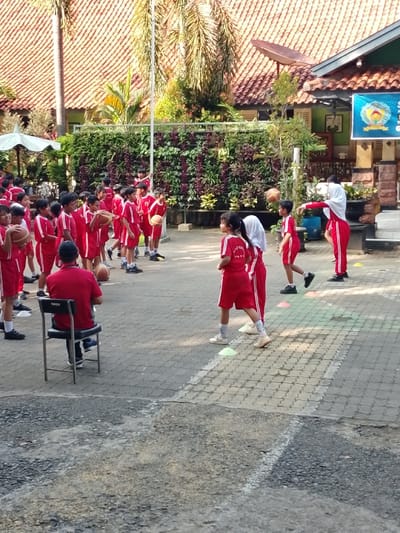 A group of children, all wearing red shirts and white shorts, are participating in a physical education class at a school in Pati, Indonesia. The class is taking place outdoors, on a paved area with a brick pattern. Several orange cones are visible on the ground, suggesting some sort of drill or game. One child holds a basketball. Two children are being instructed by an adult male teacher, wearing a red shirt, sitting on a chair, observing. There are other children in the background, including a girl in a white hijab. There is a green wall with plants in the background, followed by an orange tiled roof.