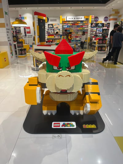 Here is a description of the image:
The image shows a life-size Lego sculpture of Bowser, the main antagonist from the Super Mario video game franchise, in a brightly lit Lego store. The sculpture is made entirely of Lego bricks and stands on a black platform with the "LEGO Super Mario" logo prominently displayed. An information card is next to the logo on the platform. In the background, the Lego store has shelves stocked with Lego sets, a "Pick & Build" section with bins of loose Lego bricks, and other displays. People are browsing the Lego products. The store appears to be located in Angelópolis, a district of Puebla, Mexico, judging by the signage on the store's wall.