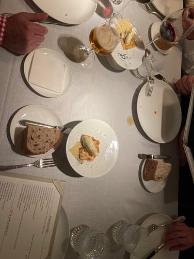 A restaurant table is set with white plates, silverware, and glasses of red wine, beer, and water. Dishes include bread and what appears to be an appetizer with a scoop of ice cream. A menu is visible, titled "Entradas Frías" and "Entradas Calientes" in Spanish, listing various cold and hot appetizers. The table is covered with a white tablecloth and has a few people around it, reading the menu.
