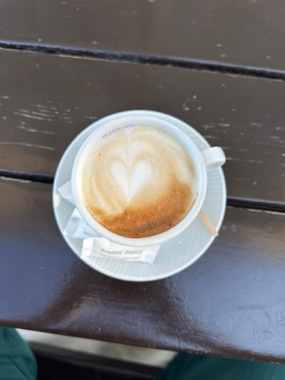 The image displays a white ceramic coffee cup containing a light brown beverage topped with frothed milk, featuring a distinct heart-shaped latte art. The cup is situated on a white ceramic saucer with a subtle ribbed texture. On the saucer, next to the cup, are two white sugar packets and a small wooden stirring stick. One sugar packet prominently displays the text "šetalište Šušanj" and "bijeli šećer 1 white sugar," indicating the location of Šušanj, Montenegro. The cup, saucer, and accompanying items are positioned on a dark brown wooden table characterized by a visible horizontal seam and scattered light reflections. In the bottom left and right corners of the frame, blurred portions of dark green fabric are visible, suggesting the presence of a seated individual.