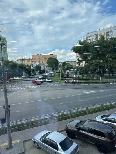 The image shows a view from a window looking out at a city street in Anapa, Russia. A busy intersection with multiple lanes of traffic is visible, with cars moving in various directions. Pedestrian crossings are marked on the asphalt. Several multi-story apartment buildings line the street, with trees and green spaces interspersed. The sky is cloudy. In the foreground, a silver sedan and a dark brown SUV are parked on a paved area next to the sidewalk. A lamppost with a trash receptacle is also in the foreground. The scene appears to be a typical urban environment during the daytime.