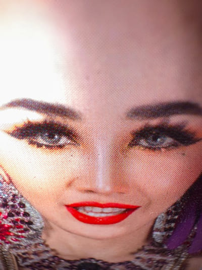This is a close-up, low-angle, and slightly distorted image of a person's face, likely a doll or a heavily made-up individual. The subject's face is turned to the side, with their left eye and mouth prominently displayed. Their makeup is striking: dark eyeliner, blue eyeshadow, and vibrant red lipstick. The skin appears smooth and has a light tone.

The background is a mix of textures and colors. Part of the subject's head is adorned with a purple headband or scarf, and some of their clothing or accessory, which has intricate patterns of black, grey, and red, is visible. The lighting is bright, creating a warm glow on the skin and a slight overexposure on the left side of the image. The image has a distinct textured quality, as if it was captured on a fabric surface or a screen with visible pixelation. The overall mood is somewhat artificial and stylized due to the strong makeup and the peculiar texture of the image. No text is visible in the image.