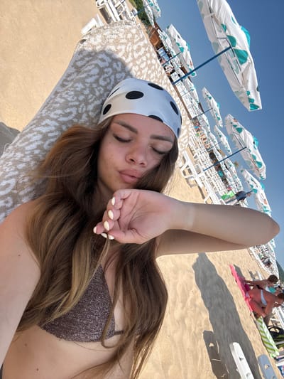 The image shows a young woman with long, brown hair lying on a towel on a sandy beach. She is wearing a white bandana with black polka dots and a sparkly brown bikini top. Her eyes are closed, and her hand is raised near her face. In the background, rows of white lounge chairs and closed beach umbrellas are visible under a clear blue sky. Some people are sitting on the beach and in lounge chairs in the distance. The scene is set on a sunny day on the beach in Primorsko, Bulgaria.