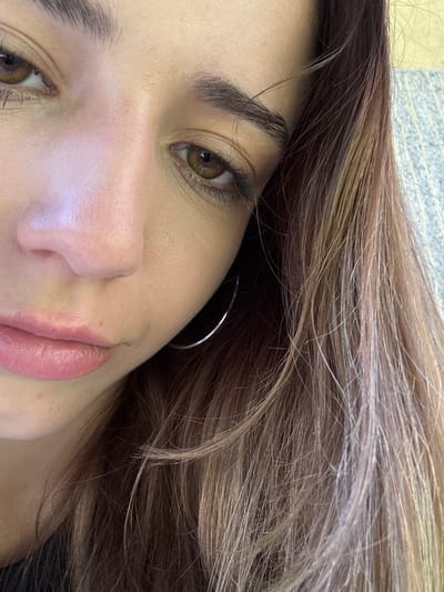 The image is a close-up, angled portrait of a young woman's face, with a focus on her eyes, nose, and lips. Her eyes are brown, with dark eyelashes and well-defined eyebrows. Her skin appears to be fair and smooth. She has light brown hair with some lighter highlights, which is partially covering her face. A silver hoop earring is visible on her left ear. The background is blurred and appears to be an indoor setting with a textured surface, possibly a sofa or wall. There are no discernible elements in the image that would indicate a location in the city of Pernik, Bulgaria, or any specific environmental context. The image is well-lit, with a soft focus.