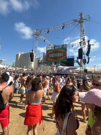 This is a wide shot of an outdoor festival or concert in Tel Aviv, Israel, on a sunny day. A large crowd of people are gathered, facing a stage setup with a large LED screen displaying the word "GENISH" in an artistic font. The screen is framed by a metal truss structure holding speakers and lighting equipment. In the background, tall buildings are visible, characteristic of the Tel Aviv skyline.
The crowd is diverse, with people of various ages and genders. Many are dressed in casual summer attire, and some are holding rainbow-colored items, suggesting this might be a Pride event. People are standing, dancing, and interacting with each other. The ground appears to be covered in dry grass or sand. Tents and smaller structures are set up around the perimeter of the event space. The sky is blue with scattered white clouds.