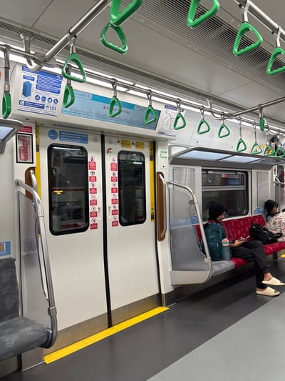 The image depicts the interior of a commuter train in East Jakarta, Indonesia. Several people are seated on the train, some appearing to rest or sleep. One person is wearing a headscarf and a backpack. Another person is wearing patterned clothing and slippers. The train carriage is equipped with numerous green safety handles for standing passengers. There are advertisements and informational posters visible on the walls, including one with the text "Layanan pelanggan 121" (Customer service 121) and another that states "Jaga barang bawaan Anda saat di dalam kereta" (Keep your belongings with you while on the train). The seats are a red patterned fabric. The lighting suggests it might be daytime, but no specific time of day is evident. The overall atmosphere appears to be calm and routine, characteristic of daily public transport.