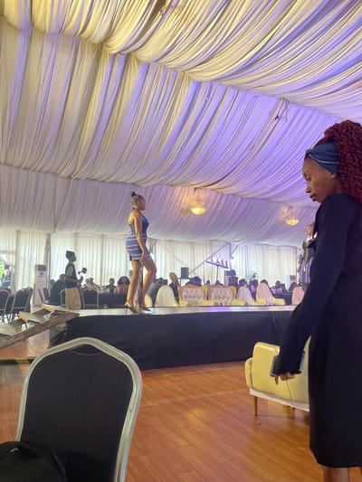 In an indoor event space in Barakin Lerawi, Nigeria, a fashion show is underway. A young woman is walking on a raised platform, modeling a short, blue and red striped dress. She has her hair styled in a high bun and wears flat, open-toed shoes. Behind her, an audience is seated at tables, with some individuals standing. Another model can be seen further down the runway. The ceiling of the venue is draped with white fabric, creating a tent-like effect, and illuminated by soft purple and yellow lights. In the foreground, a woman with curly red hair, wearing a black dress and a blue headscarf, is standing and looking towards the stage. A black chair is in the lower left of the frame, and a yellow sofa is visible on the right. The floor is polished wood.