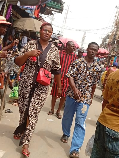 The photograph shows people walking through an outdoor market. A woman wearing a leopard print dress and red purse is in the foreground. Behind her is a man in a red and black striped shirt and another in a patterned shirt and blue sweatpants. The market stalls have umbrellas and various goods for sale. Other people are moving throughout the crowded market scene.