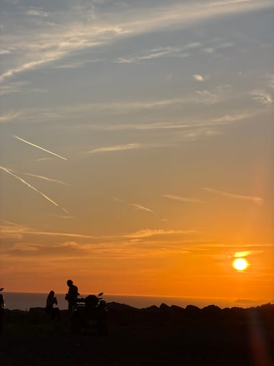 The image depicts a sunset scene in Kovačko Polje, Montenegro. In the foreground, silhouetted against the vibrant orange sky, are two people and a quad bike. One person is seated on a rocky outcrop, while the other stands beside them. The quad bike is parked to the right of the figures. The background features the sea meeting a distant, hazy coastline. The sky is adorned with wispy clouds and several distinct contrails from aircraft, streaking across the gradient of orange and blue hues. The overall impression is one of a peaceful, scenic evening in a coastal or elevated landscape.