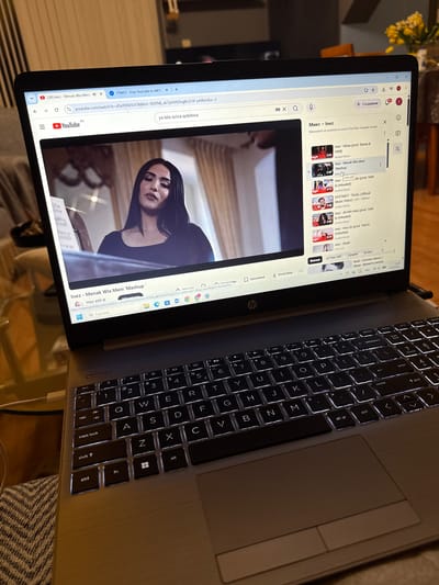 The image shows a laptop computer open and displaying a YouTube video of a woman, likely a singer or performer, in what appears to be a music video. The video is in the center of the screen, with the YouTube interface visible around it. On the right side of the screen, a list of related videos is displayed, with titles such as "Inez - Hilwa", "Inez - Menak Wla Meni 'Mashup'", and "Inez - Shabi". The YouTube page is in Bulgarian, indicated by the text "Микс - Inez" (Mix - Inez) and other Bulgarian phrases.

The laptop is placed on a surface that appears to be a couch with a patterned throw blanket. The keyboard of the laptop is illuminated, suggesting it might be dark in the room or the backlighting is turned on. The overall environment suggests a casual setting, likely indoors.

The image is a close-up of the laptop and its immediate surroundings. There is no indication of the specific location within the city of Pazardzhik, Bulgaria. The focus is on the technology and the content being viewed on the screen.