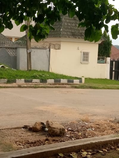 The image shows a street scene in Gwarinpa, Nigeria. In the foreground, there is a paved road with some debris and rocks on the side. A concrete curb separates the road from a grassy area. Behind the grass, there is a fence topped with barbed wire. Adjacent to the fence is a light-colored building with a grey tiled roof and a small window. A utility pole stands near the fence, with a sign attached to it. In the background, other buildings and trees are visible. The overall scene depicts a residential or commercial area in Gwarinpa.