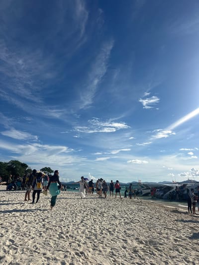 The image depicts a vibrant scene on a sandy beach, likely in Phru Nai, Thailand. The sky is a brilliant blue, scattered with wispy white clouds, suggesting a clear and sunny day, possibly mid-morning or early afternoon given the bright sunlight.
A group of people are gathered along the shoreline, with a speed boat moored nearby. Some individuals are standing, while others appear to be in the process of boarding or disembarking from the boat. Many are dressed in casual, beach-appropriate attire. The mood seems relaxed and perhaps excited, typical of a tourist excursion or day trip.
The beach itself is characterized by fine, light-colored sand, with subtle footprints indicating recent activity. In the background, lush green trees and distant hills can be seen, hinting at a tropical island setting. The water appears calm and shallow near the shore, with the speed boat indicating a mode of transport for exploring the area. Small flags, possibly national flags, are visible on the boat. There is no visible text in the image.