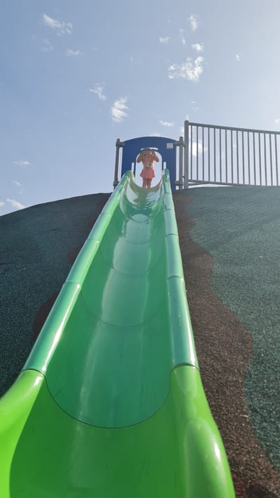A low-angle, slightly rotated, eye-level shot captures a green slide on a playground. A young child in a pink dress stands at the top of the slide, near a blue platform with a safety railing. A ladder leading to the platform is visible to the left. The playground surface is visible, showing a mix of green and brown rubberized material. The sky is blue with scattered white clouds. The playground is located in Kiryat Gat, Israel.