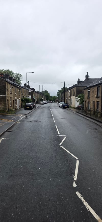 The image shows a residential street in Tameside, United Kingdom, on an overcast day. The street is wet, suggesting recent rain. Terraced houses, constructed from stone, line both sides of the road. Several cars are parked along the street and on the pavement. A road sign reading "ST MARY'S TERRACE" is visible on the left side of the image. White dashed lines mark the center of the road, with a zig-zag pattern of white lines on the right side, likely indicating a hazard or parking restriction. The sky is cloudy and grey.