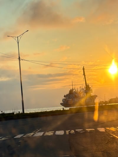 An extreme wide shot captures a sunset over the port of Petropavlovsk-Kamchatsky, Russia. The sky is awash with warm hues of orange and yellow, with clouds tinged pink and gold. A bright sunbeam pierces through the atmosphere, creating lens flare. In the middle ground, a large vessel, possibly a research or fishing ship, is docked. Its structure is complex, with cranes and multiple decks visible. The ship appears weathered. In the foreground, a road with visible white lane markings stretches across the frame, reflecting the warm light of the sunset. A tall street lamp stands to the left of the image, its silhouette stark against the vibrant sky. The sea is visible behind the ship, calm and reflecting the sky's colors. The overall atmosphere is serene, marked by the dramatic lighting of the setting sun.