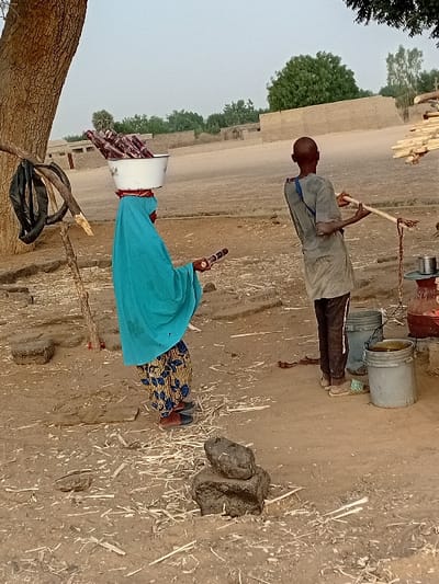 Two individuals are present in a rural landscape. A man in a light-colored shirt and dark pants operates a well system with buckets. Nearby, a woman wearing a bright blue top and patterned skirt balances a bucket filled with items on her head. A tree trunk and simple structures appear in the background. The ground is a mix of dirt, straw, and animal dung.